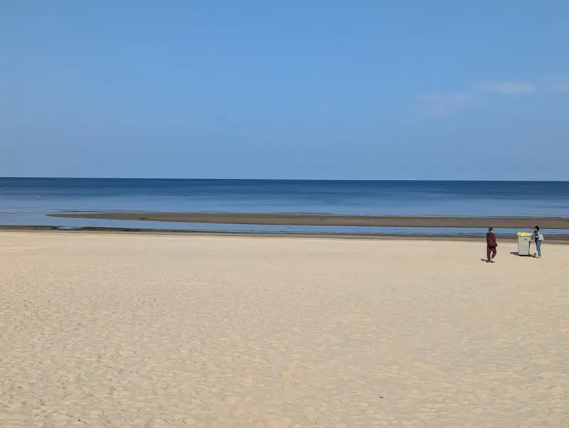 The wide flat pale-sand Baltic beach at Dzintari in Jurmala, with calm sea and a low forested shoreline